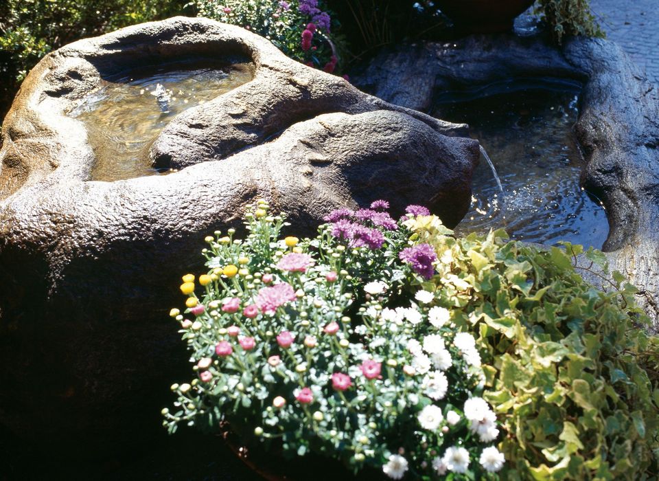 Kunstvoll geformter Steinschalen-Brunnen mit fließendem Wasser, umgeben von bunten Blumen und grünen Pflanzen