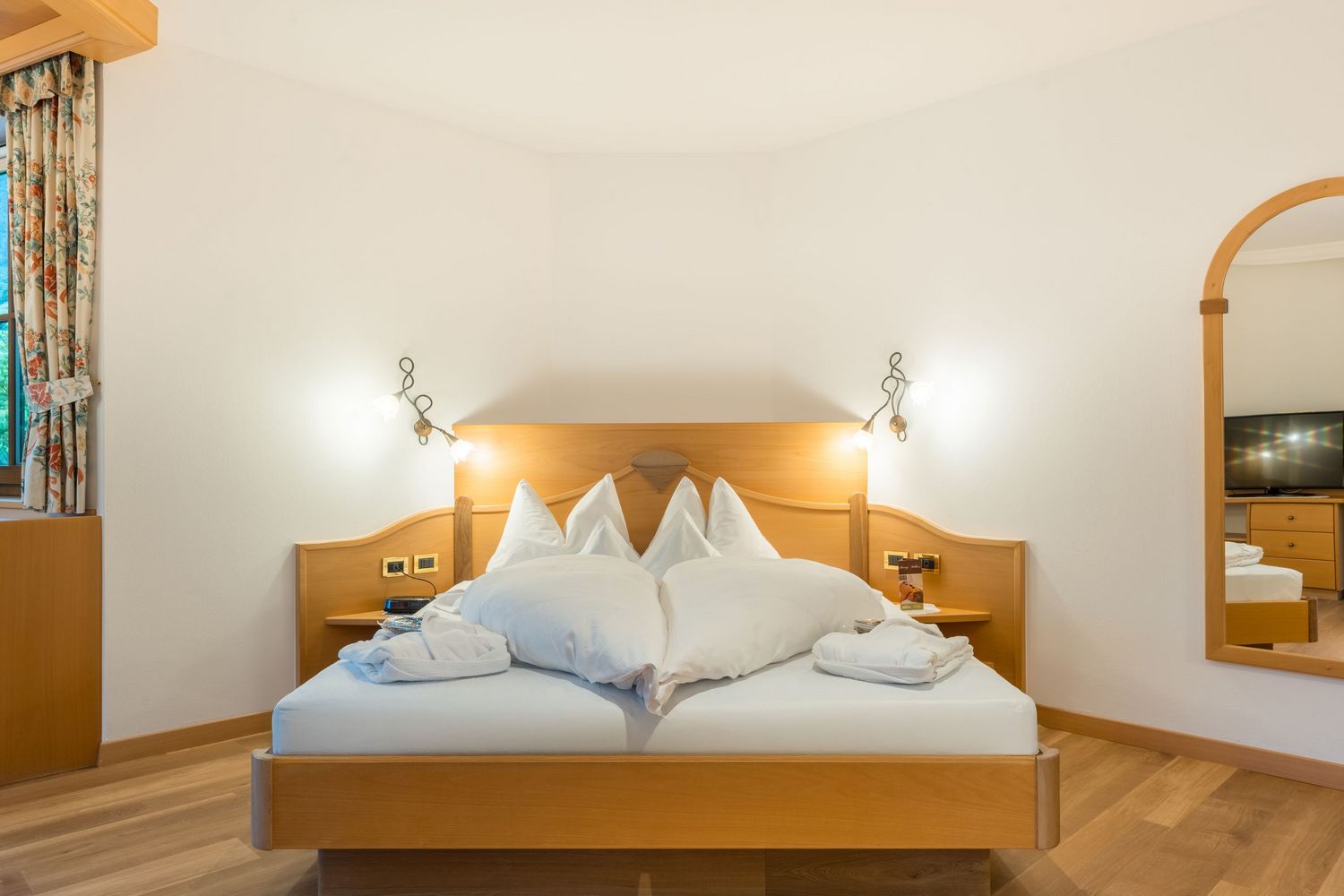 Gemütliches Hotelzimmer mit hellem Holzbett, weißen Kissen und Bettwäsche, Nachttischen mit Lampen und großem Spiegel an der Wand