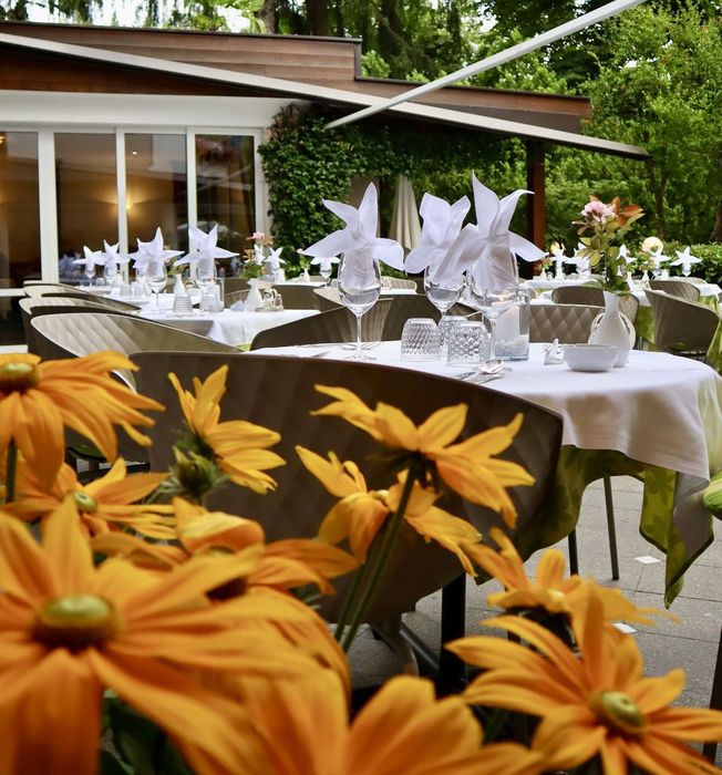 Outdoor restaurant seating with white tablecloths and floral centerpieces surrounded by green trees and yellow flowers in the foreground.