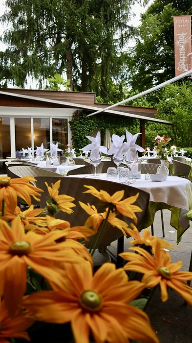 Terrassenrestaurant mit gedeckten Tischen, weißen Servietten und orangefarbenen Blumen im Vordergrund, umgeben von grünen Bäumen.