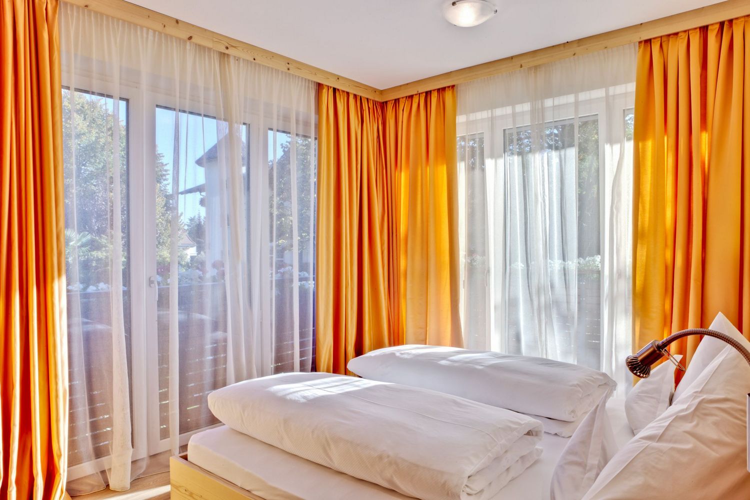 Hell eingerichtetes Schlafzimmer mit weißen Bettdecken und orangefarbenen Vorhängen vor großen Fenstern.