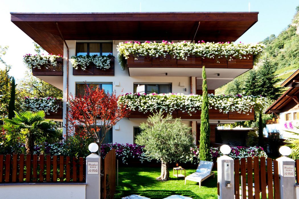Modernes Haus mit überdachten Balkonen, dekoriert mit weißen und rosa Blumen, umgeben von einem Garten mit grünen Bäumen und einer Holzlattenzaun