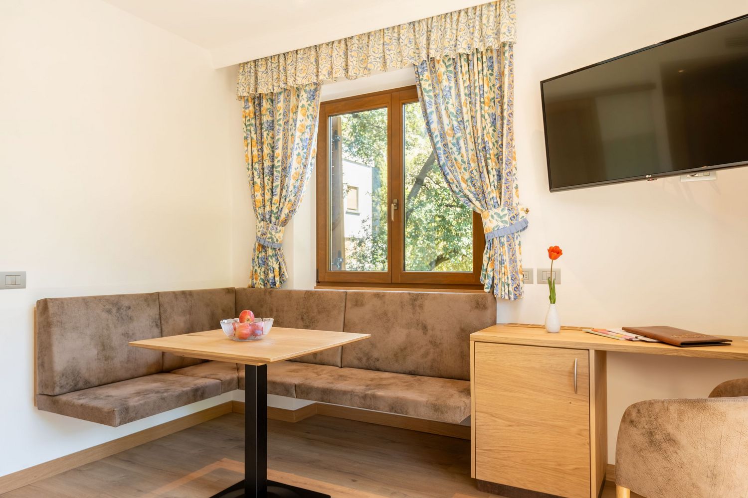 Gemütliche Sitzecke mit braunen Polstern, Holztisch, einem Fenster mit bunten Vorhängen und Fernseher an der Wand