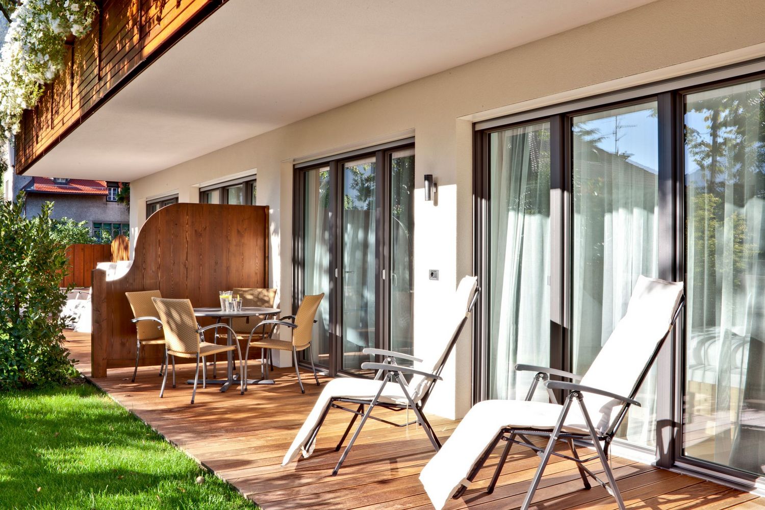 Sonnige Terrasse mit Holzdeck, zwei Liegestühlen mit weißen Auflagen und einem Tisch mit vier Stühlen vor großen Glas-Schiebetüren.