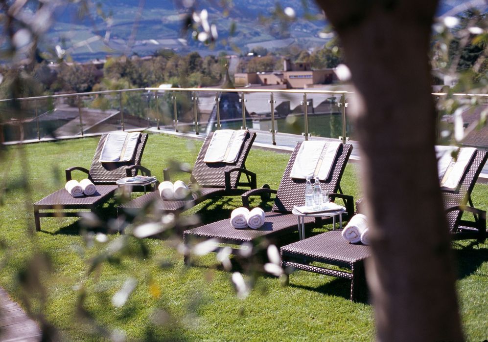 Liegestühle mit weißen Handtüchern und Wasserflaschen auf einer grünen Terrasse mit Glasgeländer und Blick auf Landschaft