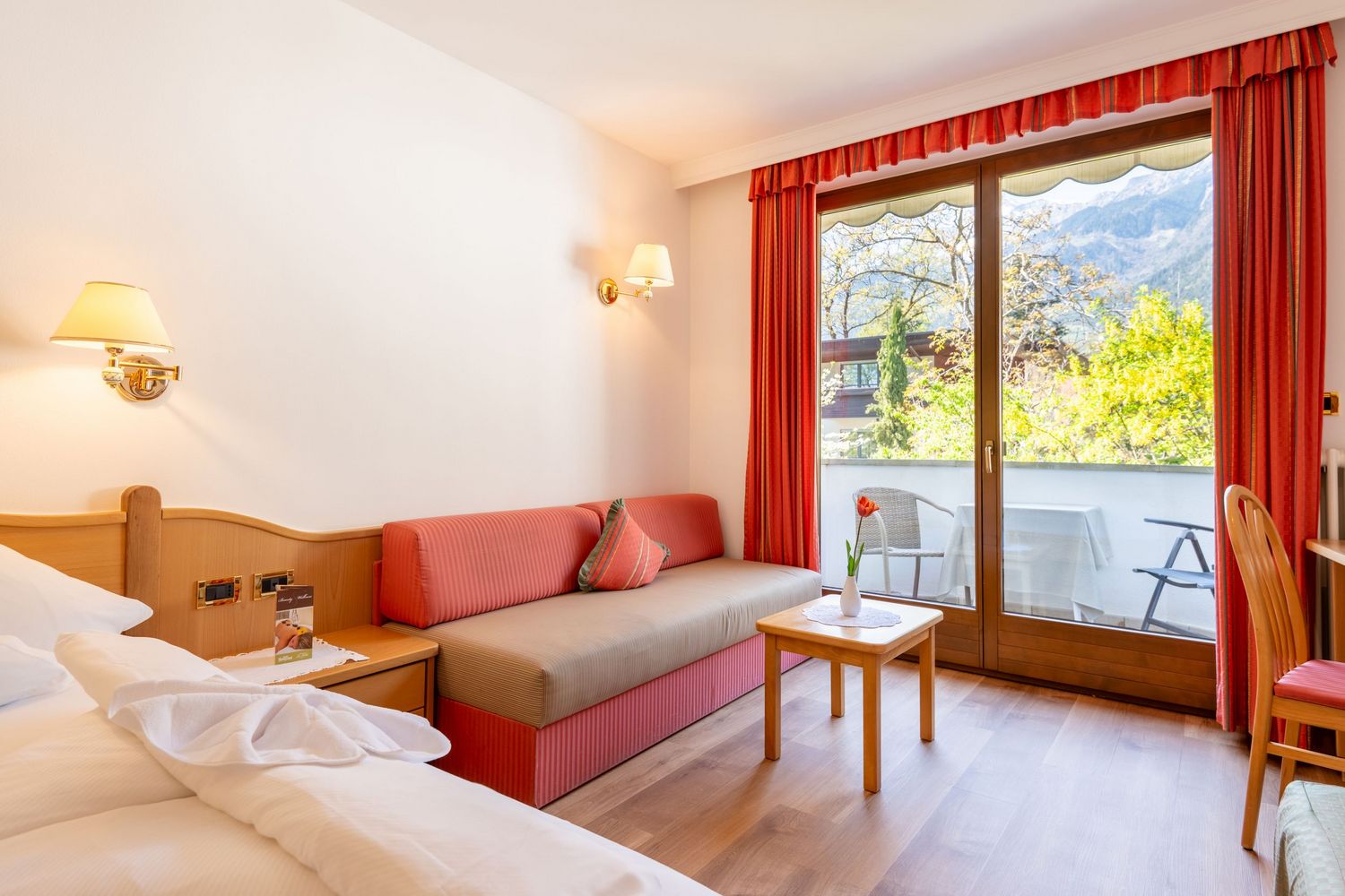 Helle Hotelzimmer mit rotem Sofa, Holzmöbeln und Blick auf Balkon mit Berglandschaft