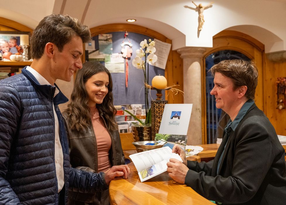 Junges Paar am Empfangstresen eines Hotels spricht mit einer Mitarbeiterin, die ein Informationsheft zeigt.