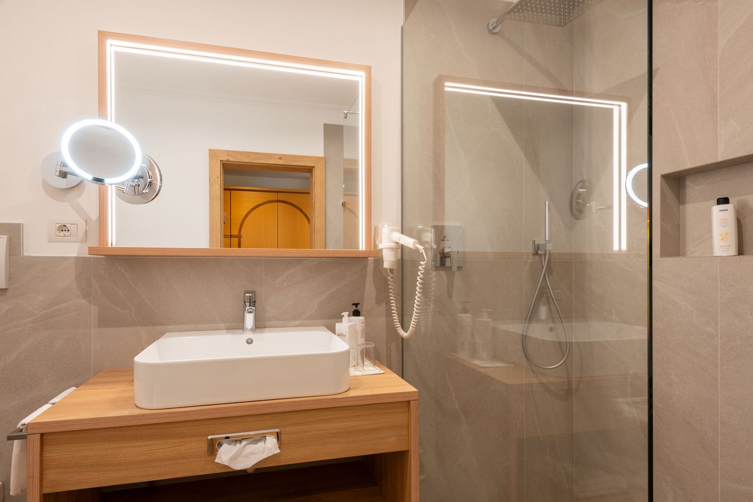 Modernes Badezimmer mit beleuchtetem Spiegel, Waschbecken auf Holzunterbau, Wandföhn und Duschbereich mit Glasabtrennung