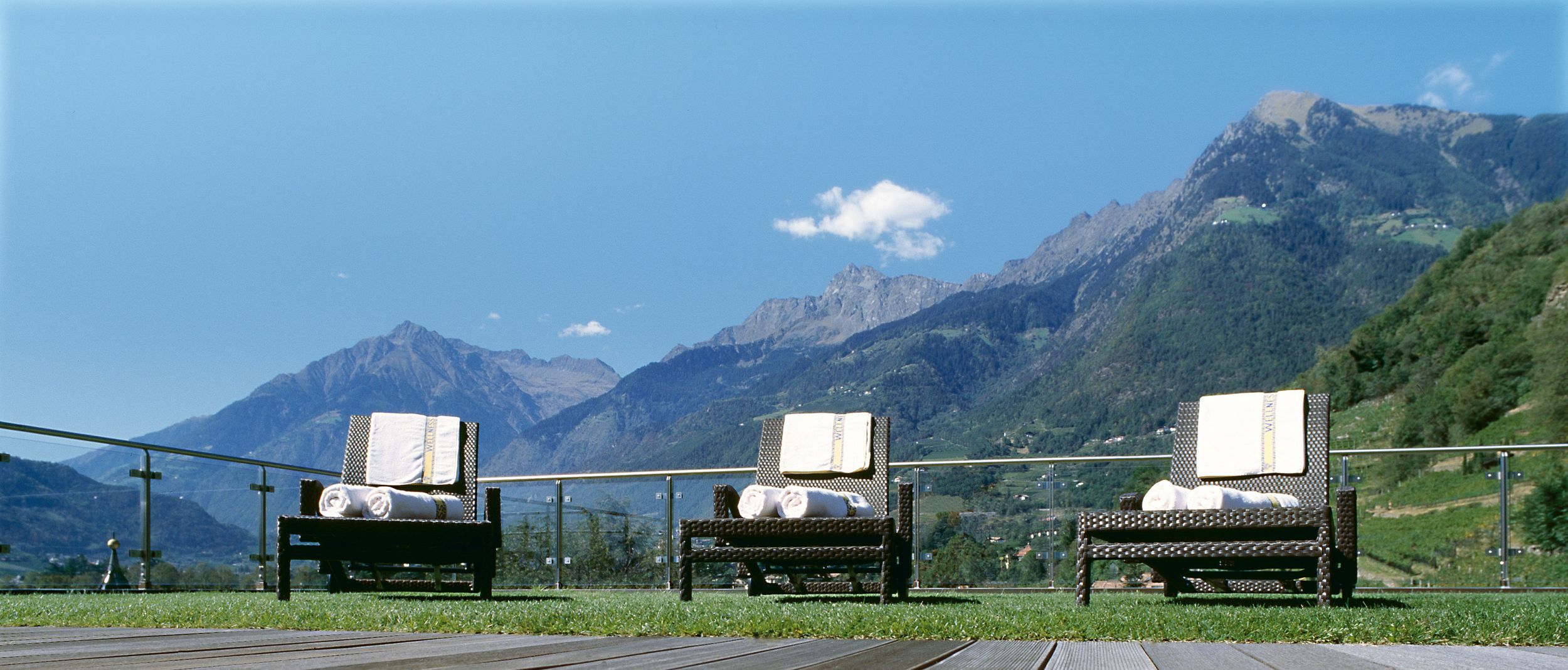 Drei Liegestühle mit weißen Handtüchern vor Bergkulisse und blauem Himmel auf Holzterrasse
