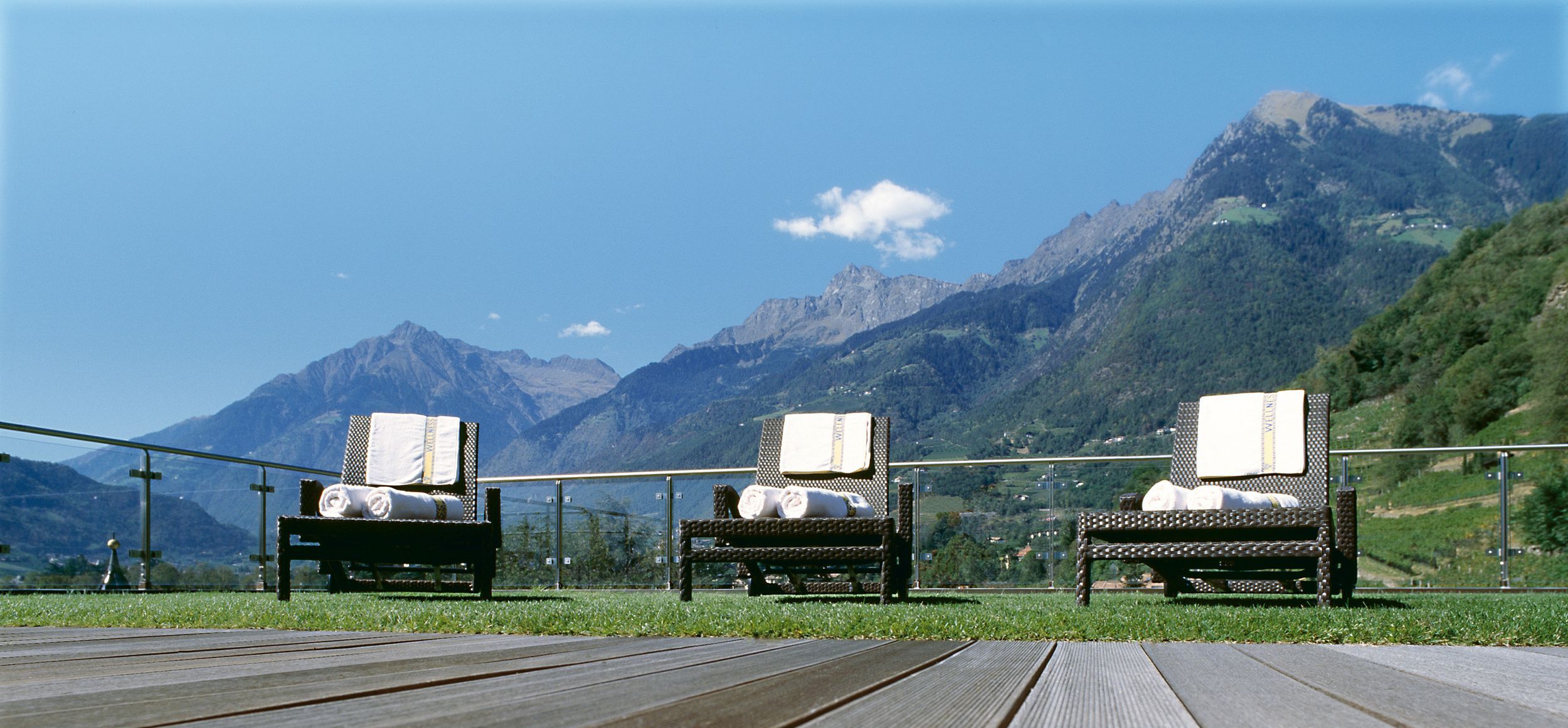 Three lounge chairs with towels on a patio overlooking mountains under a clear blue sky.