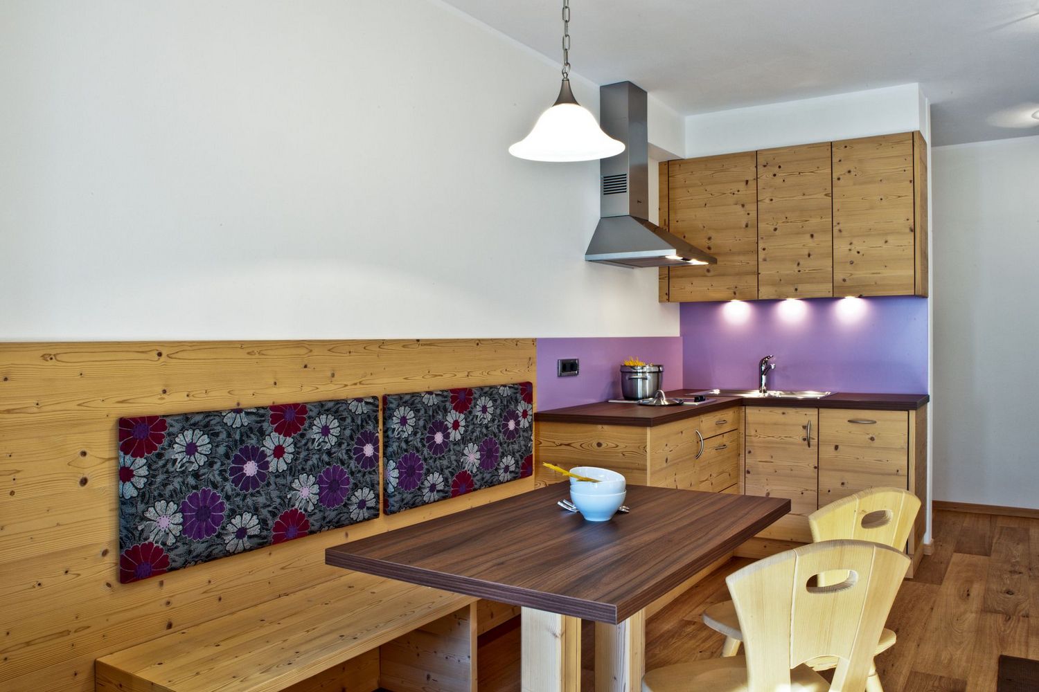 Kleine helle Küche mit Holzmöbeln, violettem Rückwandbereich und Esstisch mit gelben Stühlen.