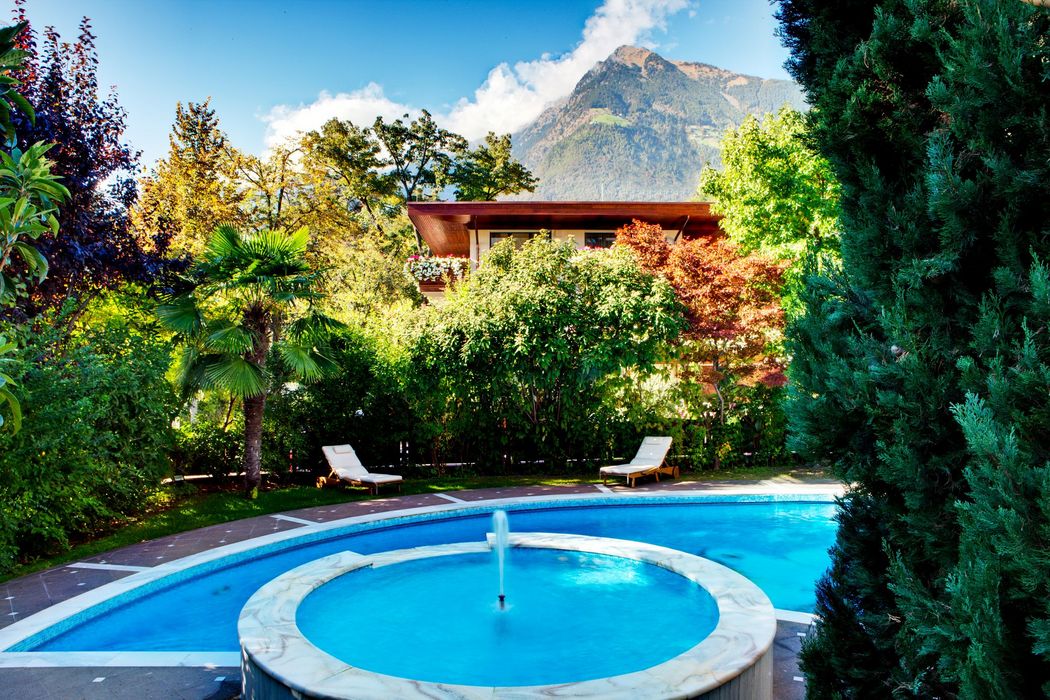 Runder Pool mit Springbrunnen, umgeben von grünen Bäumen, Liegestühlen und einem Haus im Hintergrund vor Bergkulisse