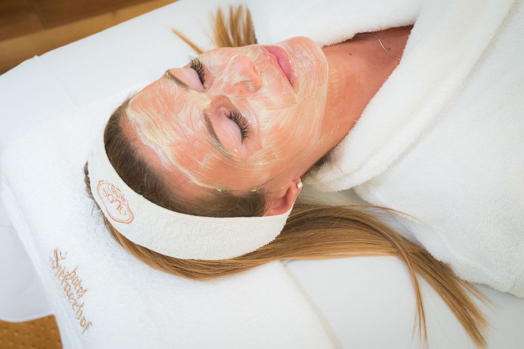 Woman with a facial mask lying on a spa bed wearing a white headband and robe.