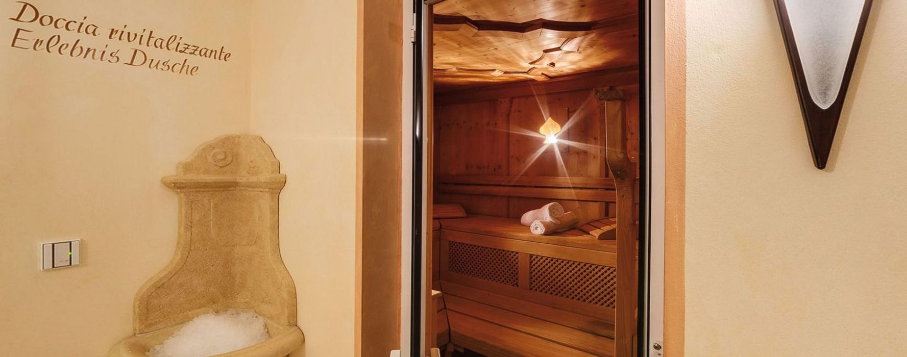 Blick in eine gemütliche Sauna mit Holzwänden und Handtüchern, daneben eine alte steinerne Wandbrunnen mit Eis und Schriftzug an der Wand