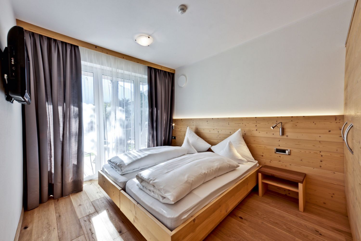 Helles Schlafzimmer mit Doppelbett aus Holz, weißen Bettdecken, einem Nachttisch, Gardinen und einem Fernseher an der Wand.