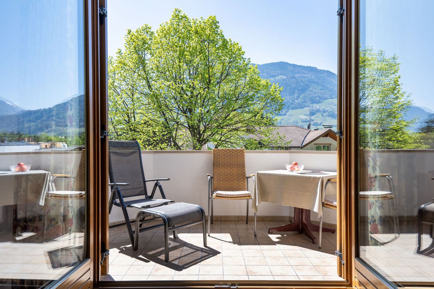 Balkon mit zwei Stühlen, ein Tisch mit Tischdecke und frischem Obst, grüner Baum und Bergblick im Hintergrund