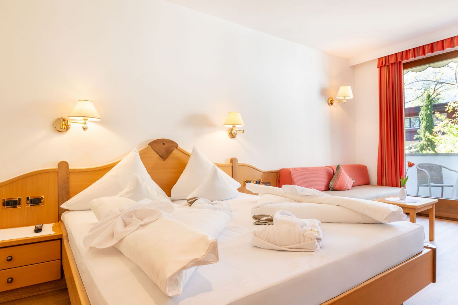 Helles Hotelzimmer mit großem Bett, weißen Kissen und Bettwäsche, rotem Sofa, Holzmöbeln und Balkon mit Ausblick auf Bäume.