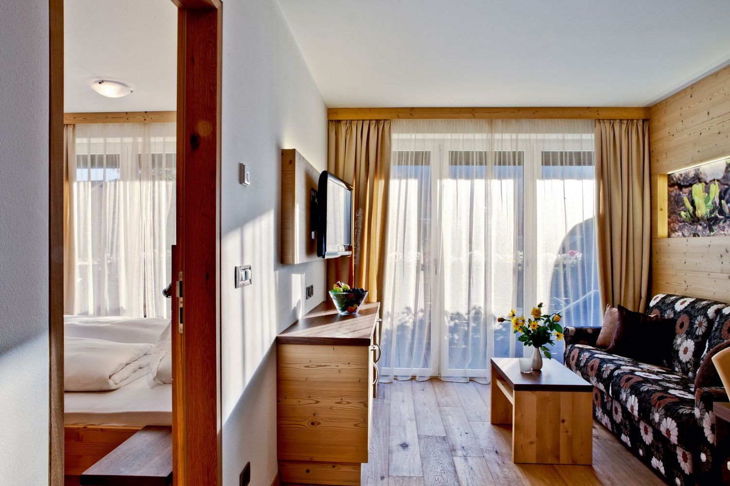 Helles Wohnzimmer mit Holzvertäfelung, floralem Sofa, Holztisch mit Blumen und angrenzendem Schlafzimmer mit Bett.