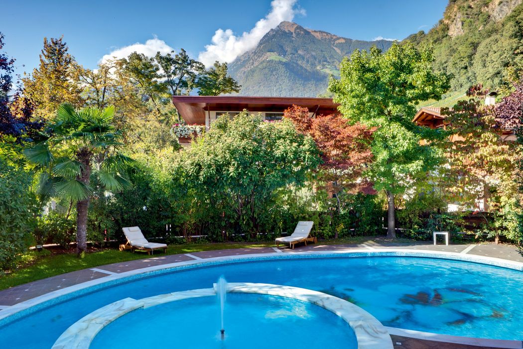 Runder Pool mit Wasserfontäne und Liegestühlen, umgeben von Bäumen und Bergen im Hintergrund