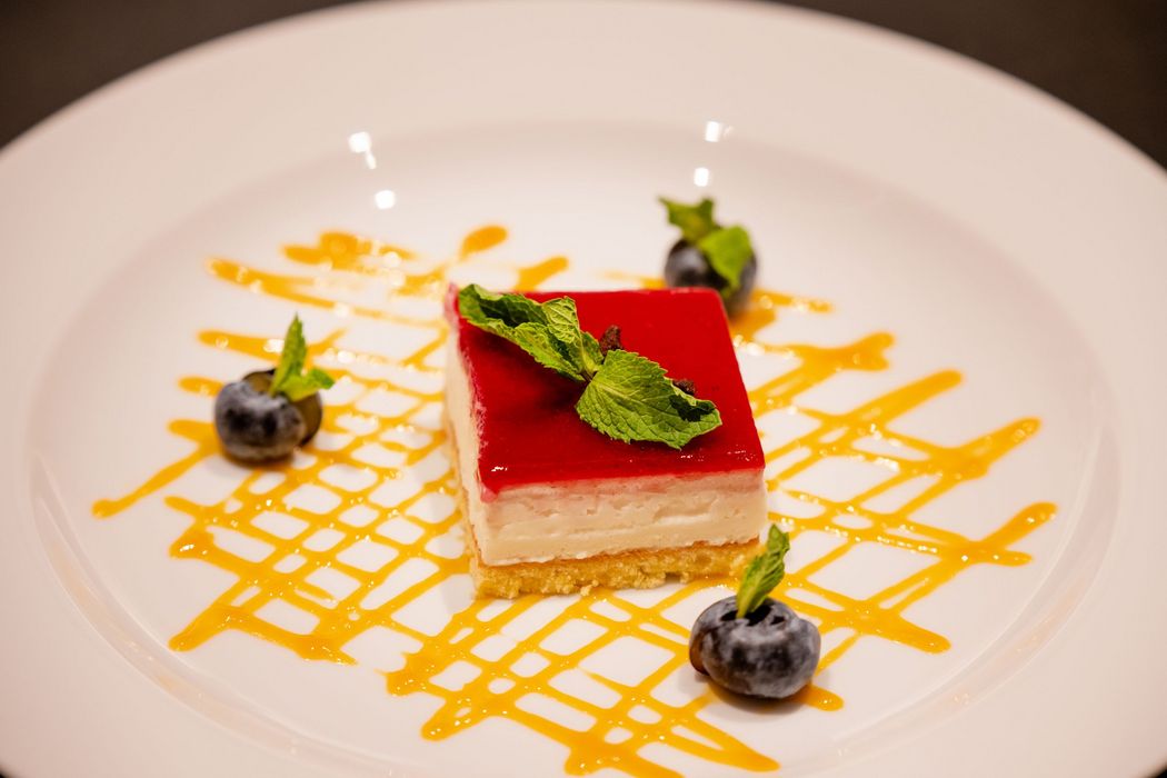 Quadratisches Dessert mit roter Glasur, Minzblättern und Blaubeeren auf weißem Teller mit gelber Soßendekoration.