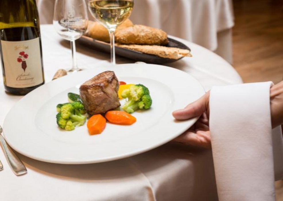 Servierperson reicht Teller mit gebratenem Fleisch, Karotten und Brokkoli in elegantem Restaurant, im Hintergrund Weinflasche, Weinglas und Brot