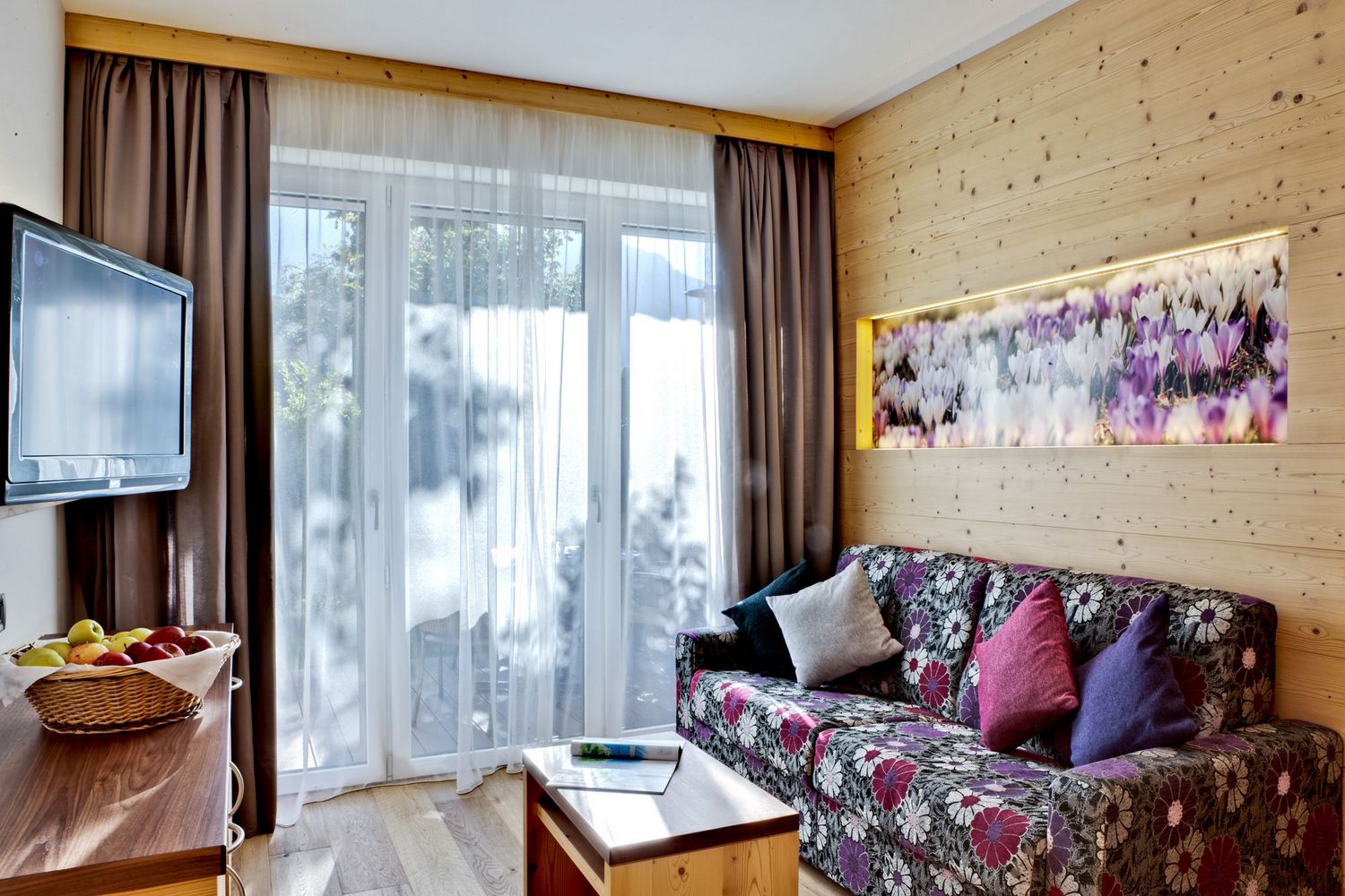 Gemütliches Wohnzimmer mit bunter Blumencouch, Kissen, Holzmöbeln und großen Fenstern mit Gardinen