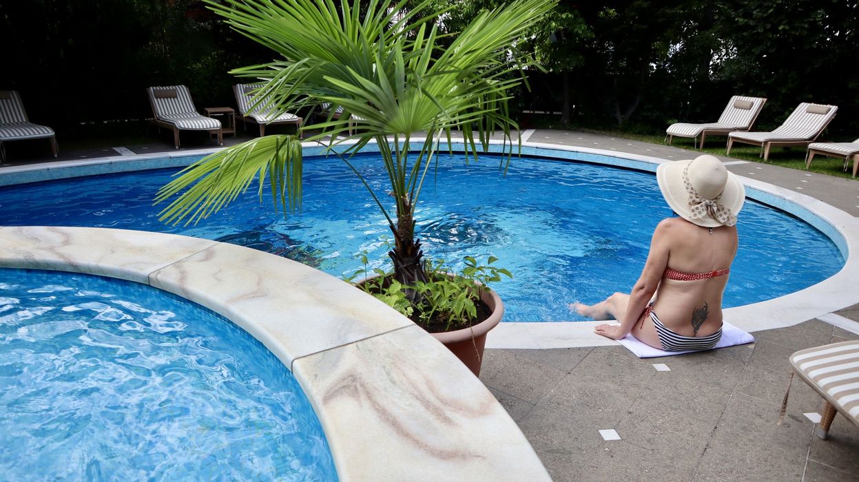 Frau im Bikini mit Sonnenhut sitzt am Rand eines runden Pools, daneben ein kleiner Pool und eine Topfpflanze