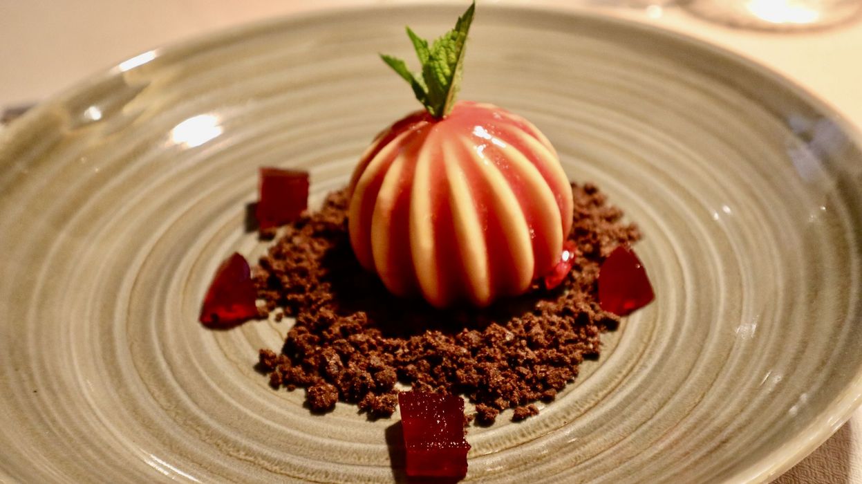 Elegantes Dessert mit einer Kugel Vanille-Eis, umgeben von Schokoladenkrümeln und roten Geleestücken auf einem grauen Teller