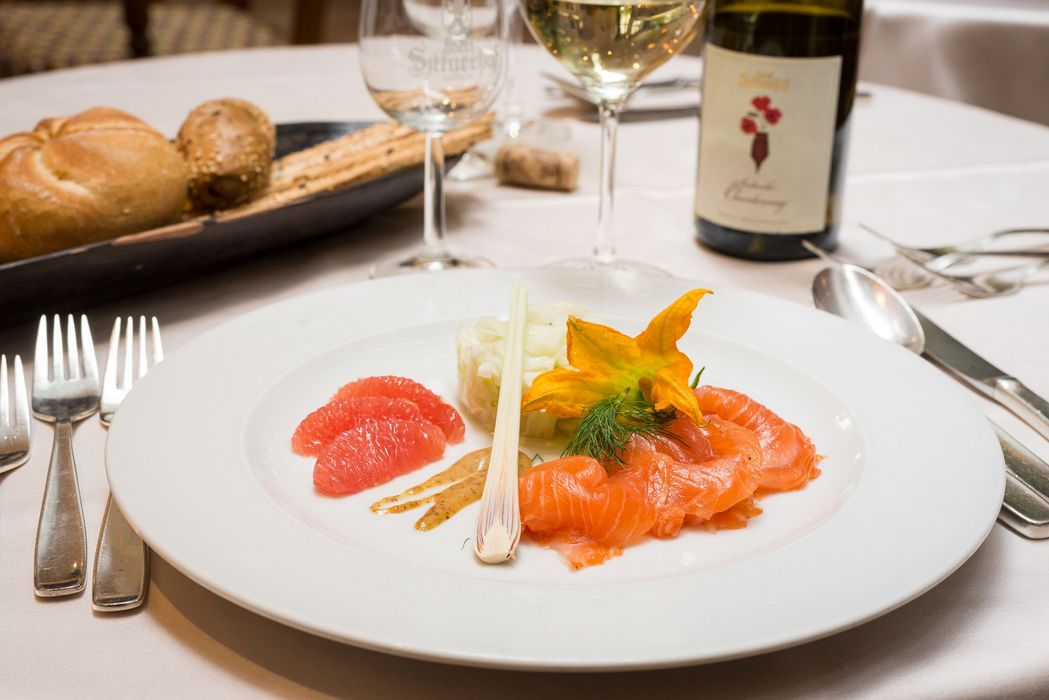 Fein angerichtete Vorspeise mit Lachs, Grapefruit, Gurkensalat und Blüte auf weißem Teller, Brotkorb und Weißwein im Hintergrund