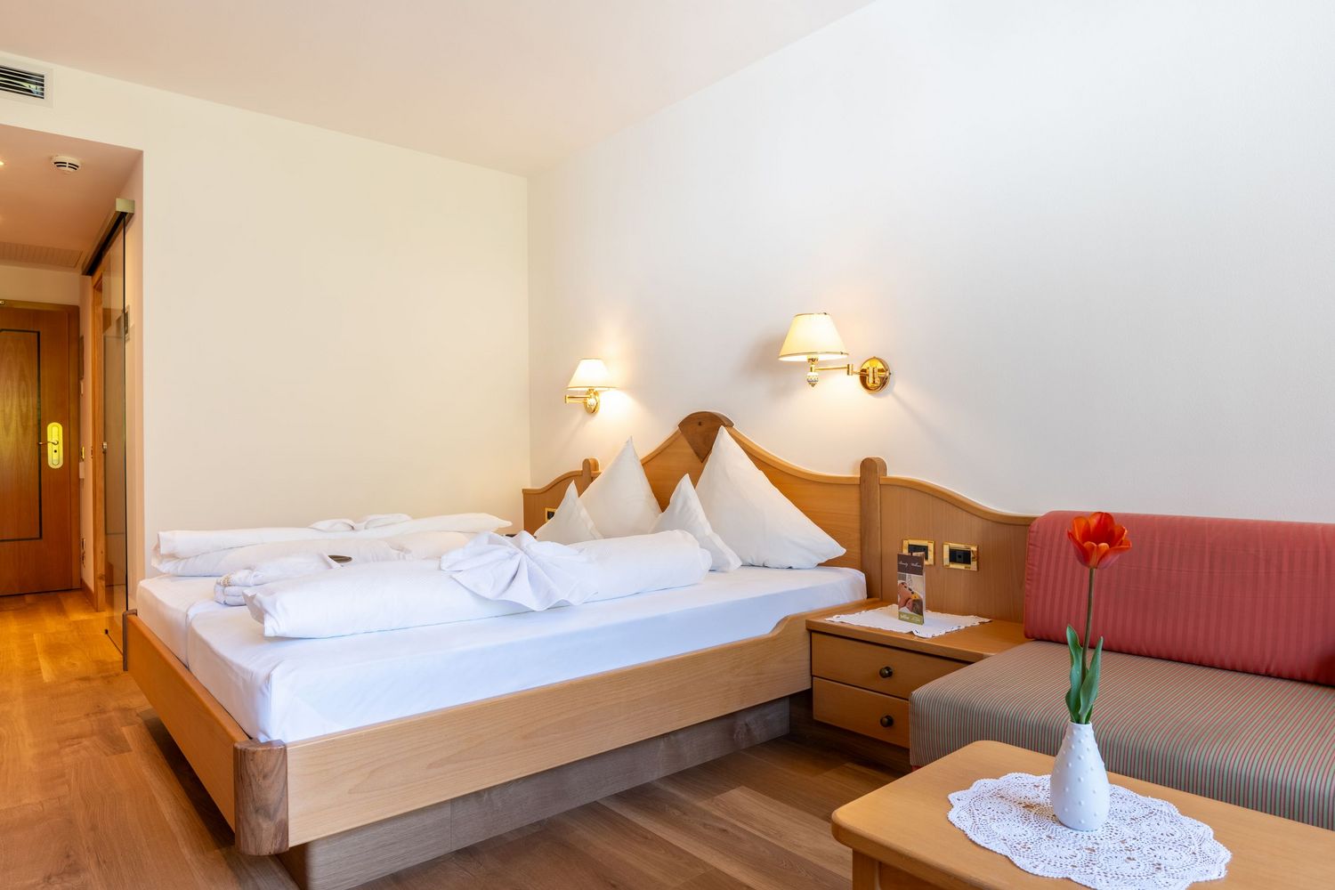 Hell eingerichtetes Hotelzimmer mit Doppelbett, weißen Kissen, Nachttisch, Wandlampen und rotem Sofa mit Blume auf dem Tisch