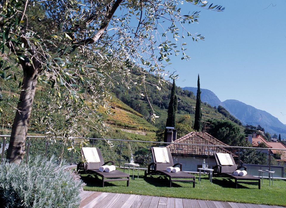 Drei Liegestühle mit weißen Handtüchern auf einer Terrasse mit Blick auf grüne Weinberge und Berge im Hintergrund