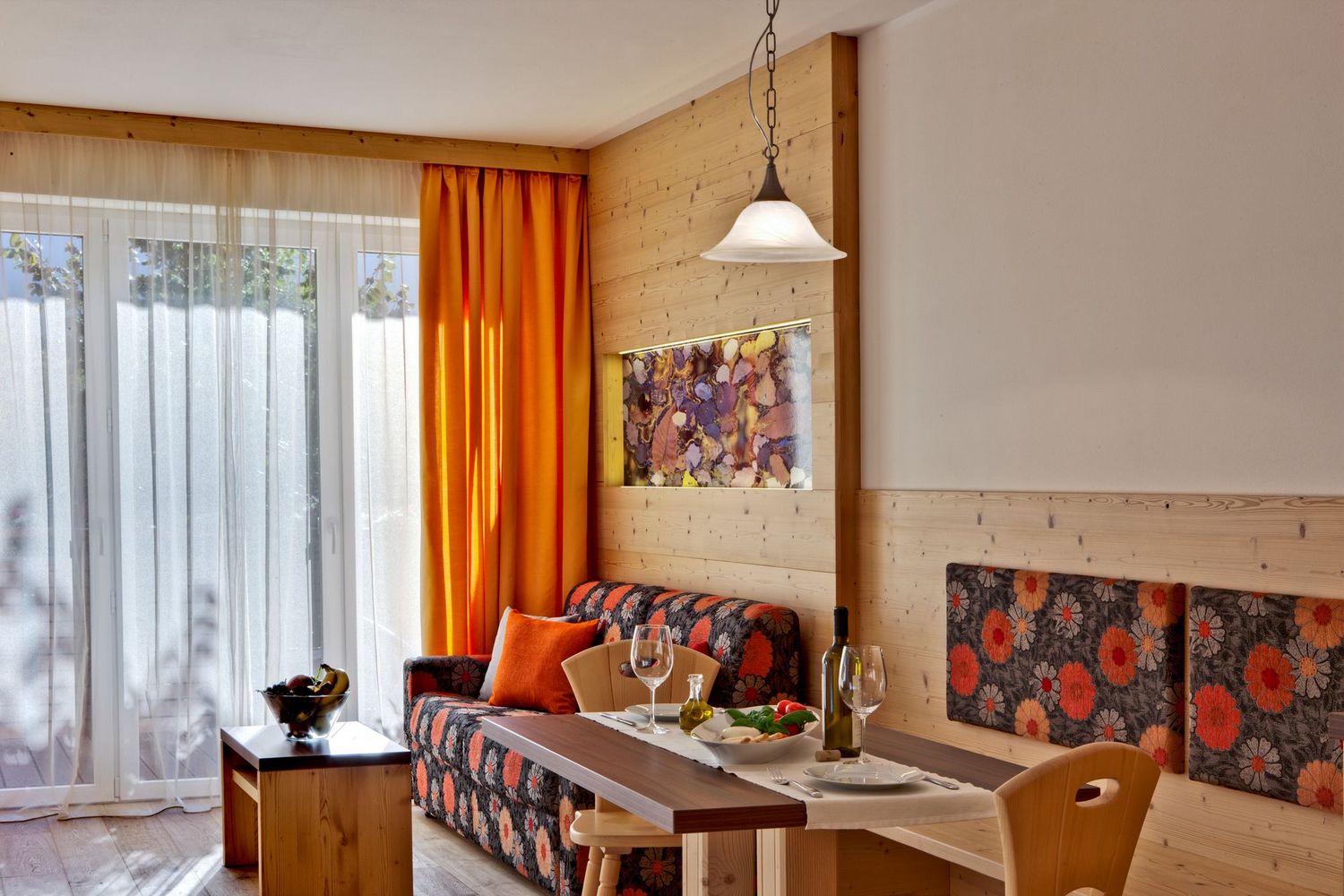 Gemütlicher Essbereich mit braun-orangen floralen Möbeln, Holztisch, orangefarbenen Gardinen und großen Fenstern mit weißen Vorhängen.
