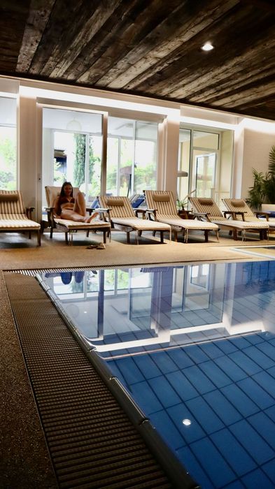 Innenbereich eines Schwimmbads mit mehreren Liegestühlen und einer Person, die auf einem Liegestuhl entspannt, reflektiert im Poolwasser