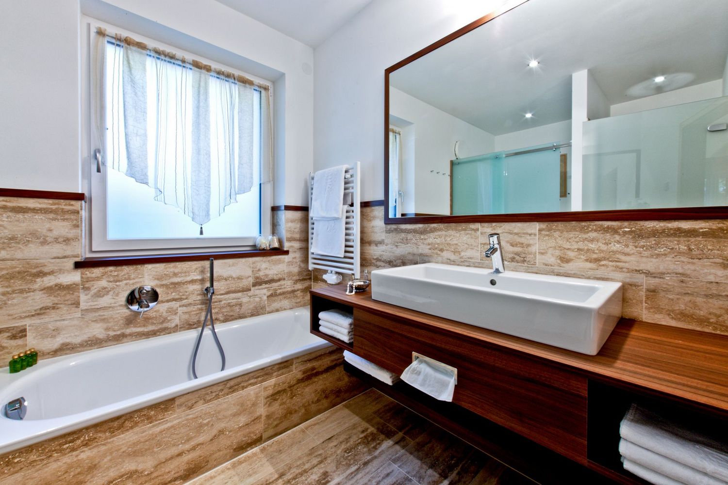 Modernes Badezimmer mit Badewanne, Waschtisch aus Holz, großem Spiegel und Fenster mit weißer Gardine