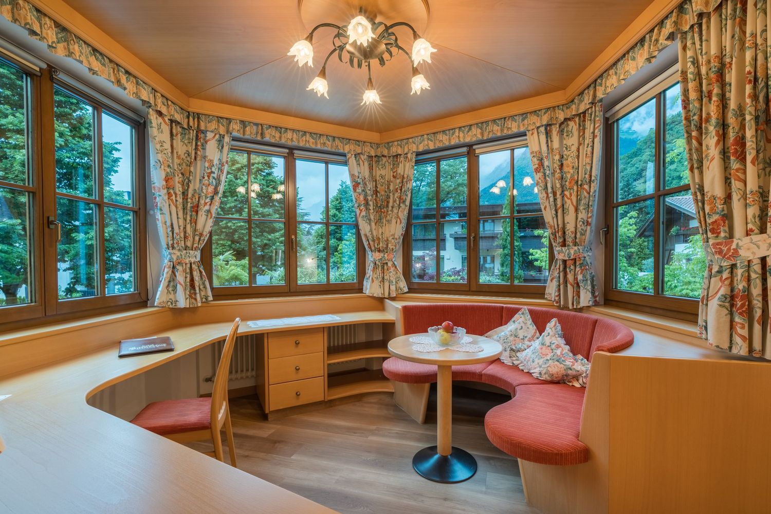 Gemütliche Essecke mit roter Polsterbank, rundem Holztisch, floral gemusterten Gardinen und großen Fenstern mit Blick ins Grüne