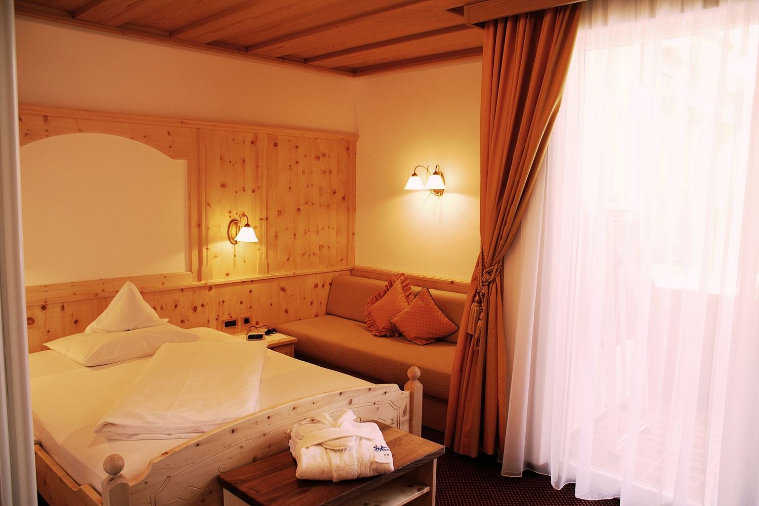Gemütliches Hotelzimmer mit Holzmöbeln, Bett, Sofa, warmem Licht und Vorhängen am Fenster