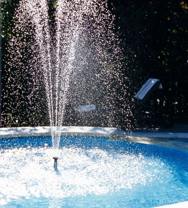 Water fountain spraying water droplets in a pool with greenery in the background.