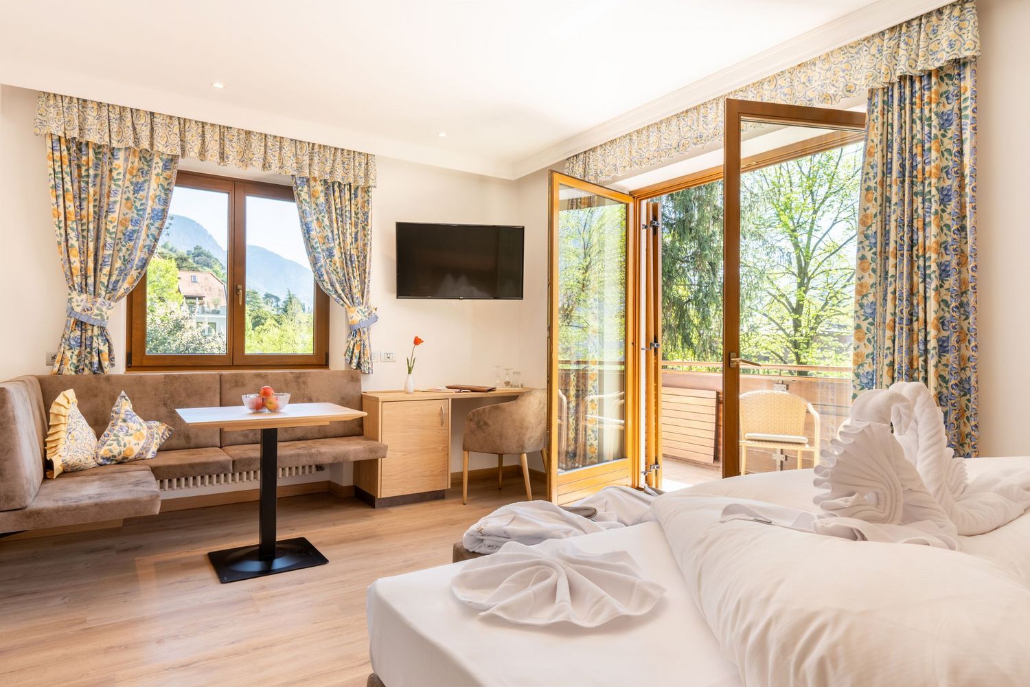 Helles Hotelzimmer mit offenem Balkon, gedecktem Tisch, Fernseher und dekorativ gefalteten Handtüchern auf dem Bett.