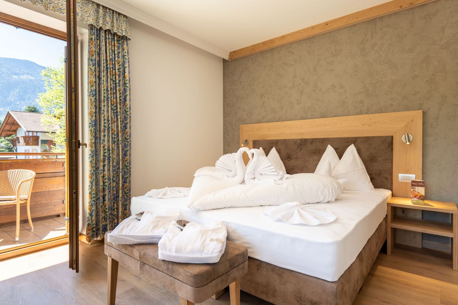 Helles Hotelzimmer mit einem großen Bett, auf dem Handtücher zu zwei Schwänen geformt sind, und einem Balkon mit Bergblick.