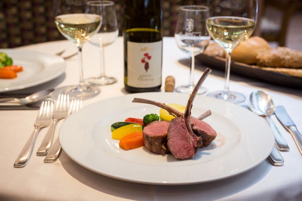 Elegantes Gericht mit Lammkoteletts, buntem Gemüse und Weißwein auf weiß gedecktem Tisch