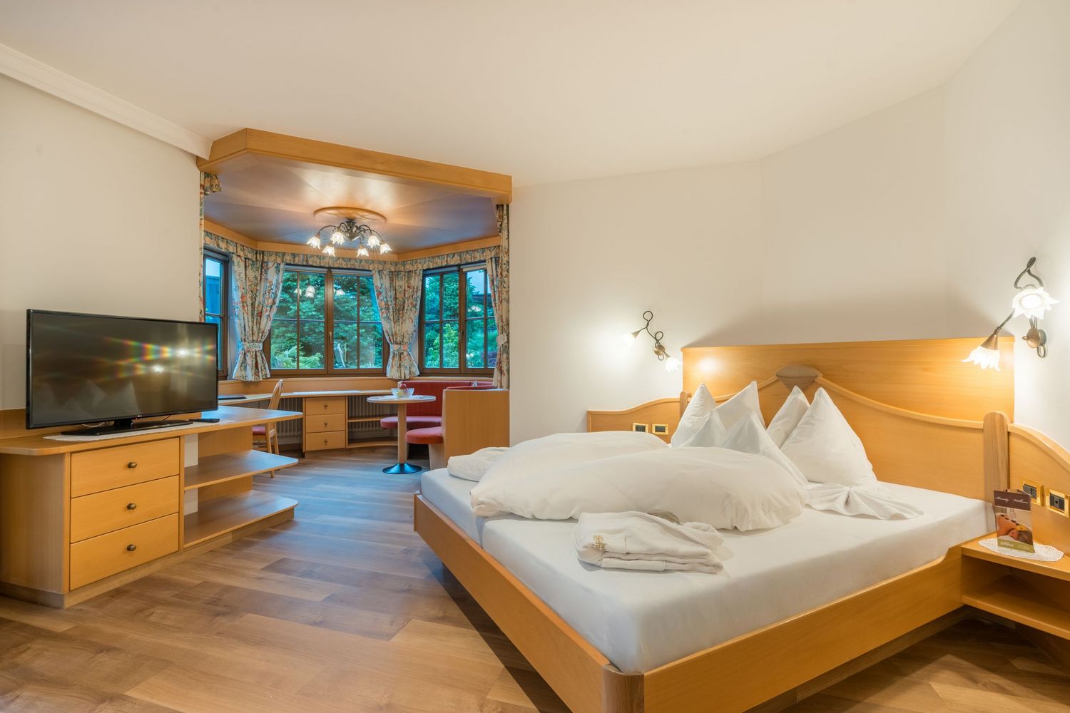 Helles Hotelzimmer mit Holzmöbeln, großem Bett, Fernseher und Sitzecke vor Fenster mit Blumenmuster-Vorhängen