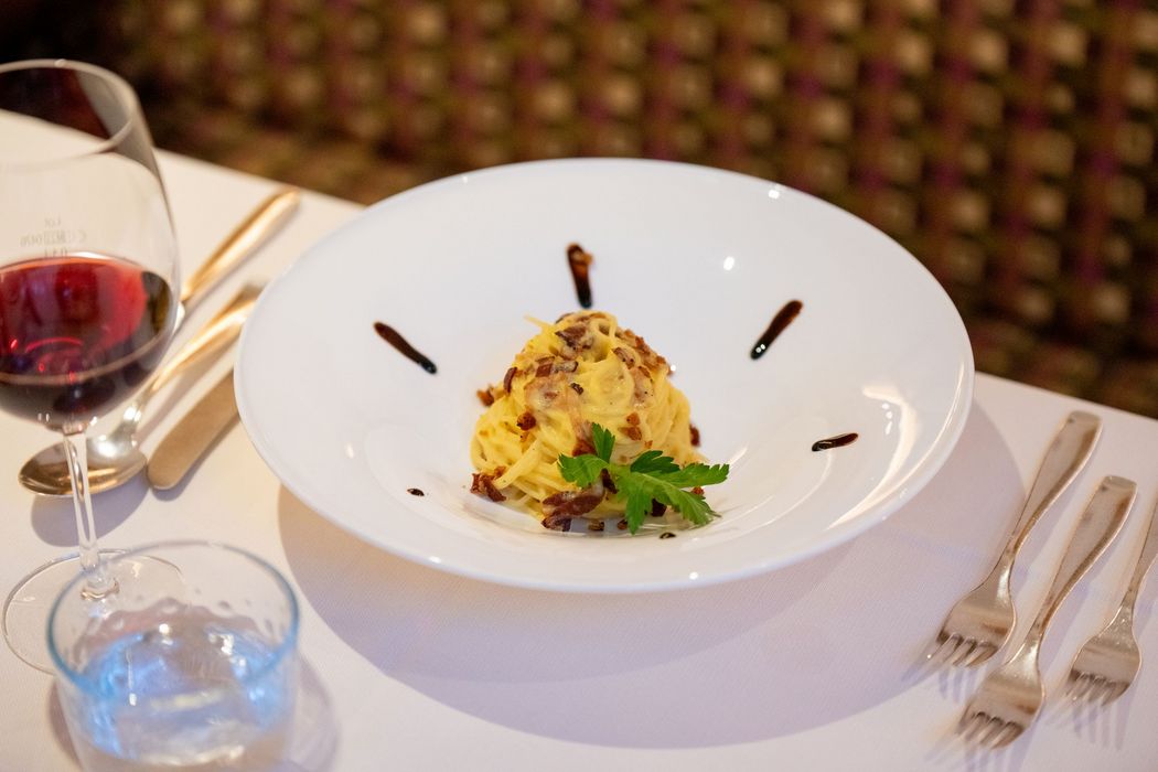 Ein Teller mit einer Portion Pasta, garniert mit Petersilie und Balsamico, neben Weinglas und Wasserglas auf gedecktem Tisch.