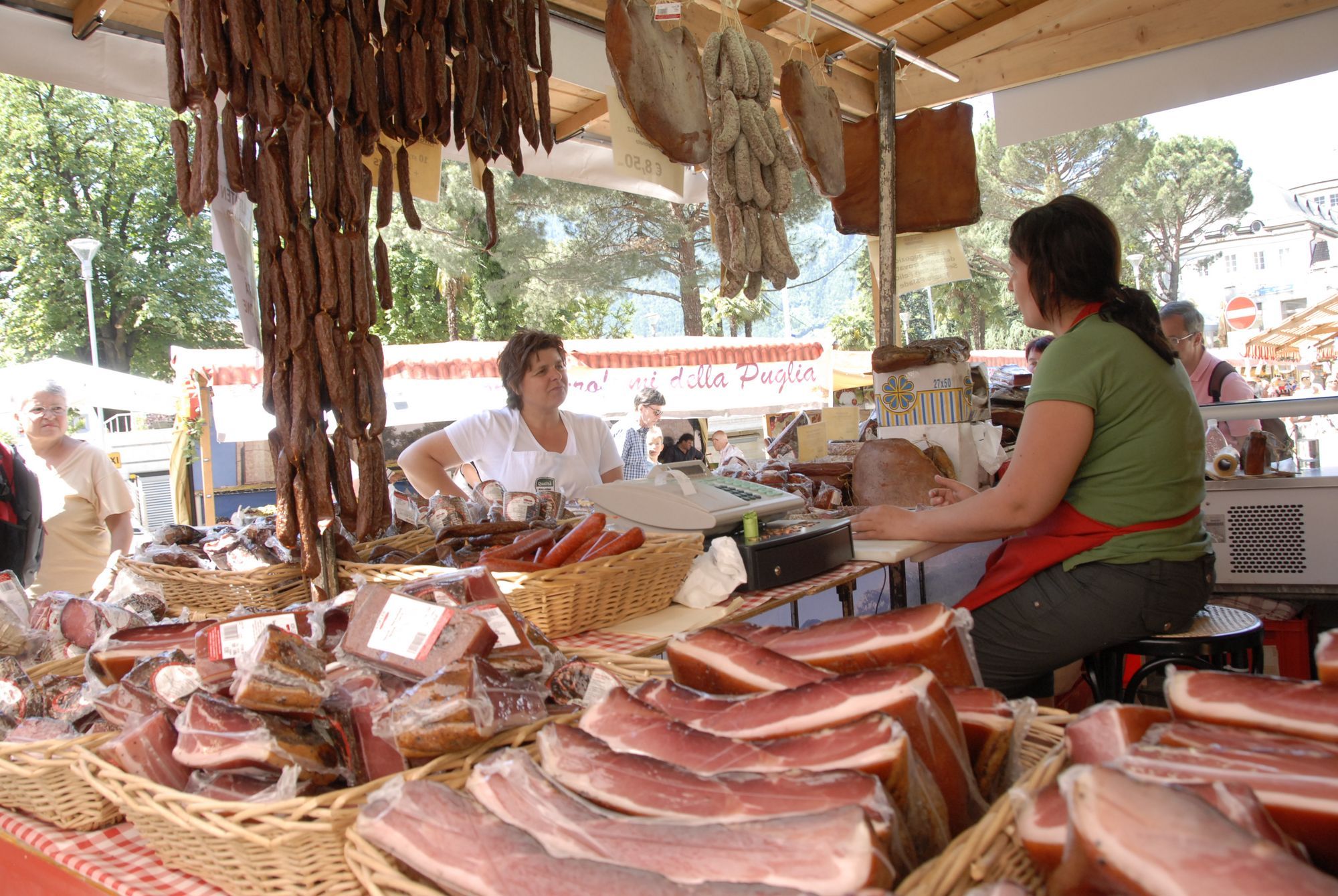 Outdoor market stall with hanging sausages and various cured meats on display, two women behind the counter.