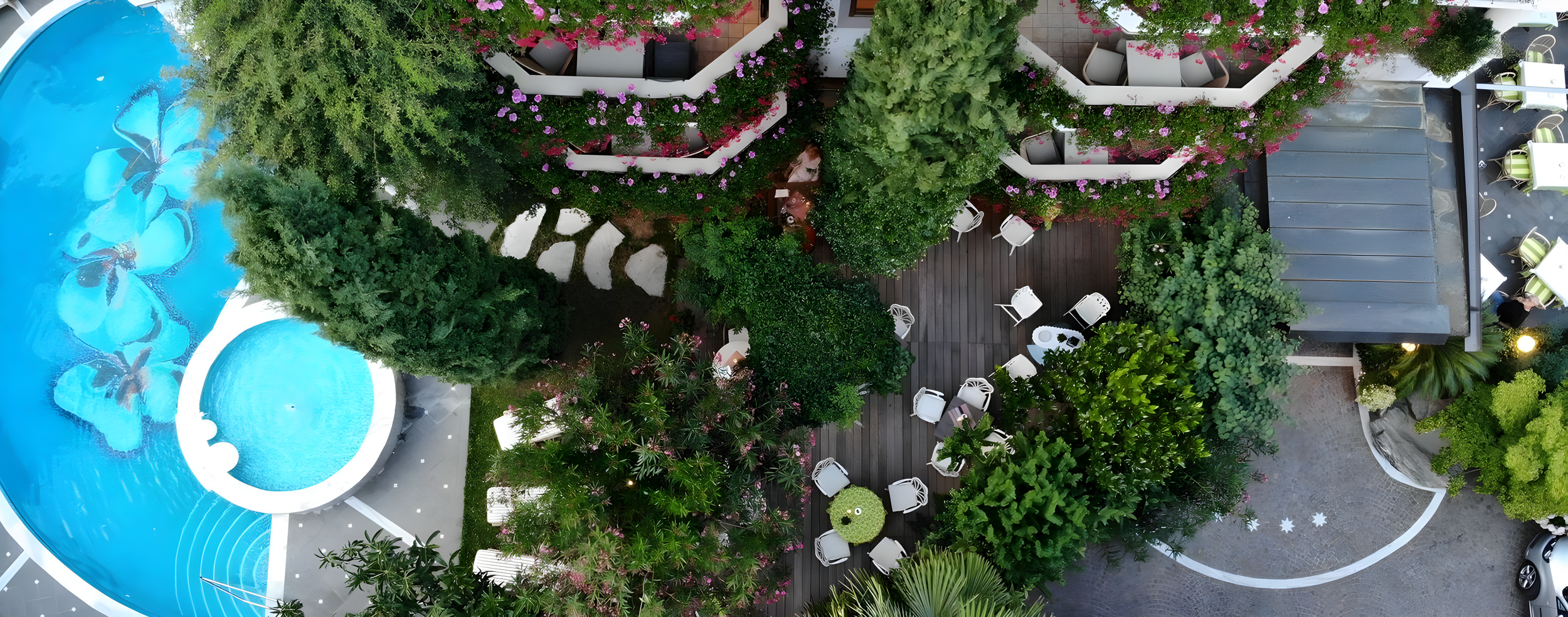 Vista aerea di un giardino con piscina decorata con fiori, tavoli e sedie all'aperto, balconi fioriti e alberi rigogliosi