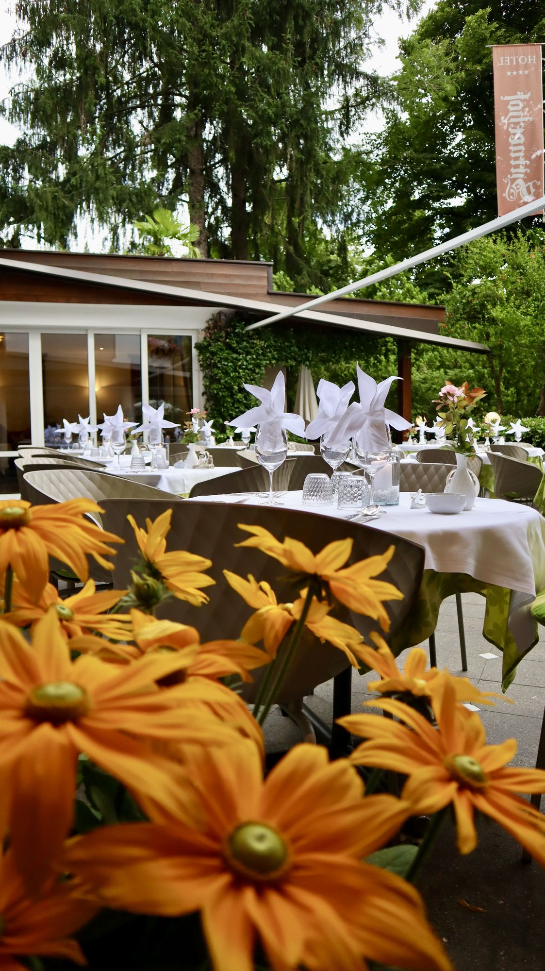 Terrazza di un ristorante elegante con tavoli apparecchiati, fiori gialli in primo piano e alberi verdi sullo sfondo