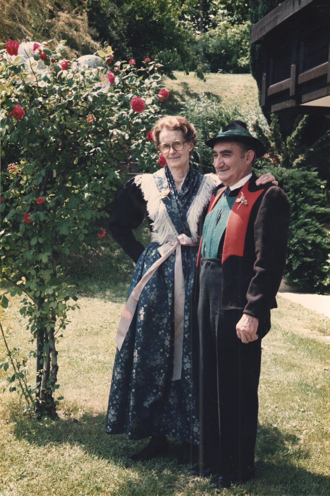 Elderly couple in traditional attire standing outdoors beside a rose bush with greenery and a house in the background.
